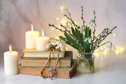 Bouquet of Snowdrop flowers with willow branches, christian rosary beads, books and candles on table, abstract blurred background. Religious church holiday. symbol of faith in God, Easter, Palm Sunday