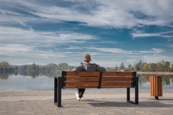 A young guy sits on a bench by the lake. A warm calm day, light and white clouds in the sky