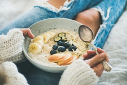 Healthy winter breakfast in bed. Woman in woolen sweater and shabby jeans eating vegan almond milk oatmeal porridge in bowl with berries, fruit and almonds. Clean eating, vegetarian food concept