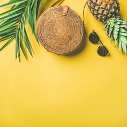 Colorful summer female fashion outfit flat-lay. Straw hat, bamboo bag, sunglasses, palm branches, pineapple over yellow background, top view, copy space, square crop. Summer fashion, holiday concept