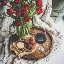 Wine snack set with flowers. Glass of red wine, cheese, roasted almonds, strawberries and bouquet of Spring red tulips on wooden tray over white knitted blanket, square crop. Romantic mood concept