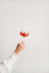 Womans hand in white shirt holding and turning glass of rose wine over white wall background. Wine shop, wine tasting, bar, wine list concept