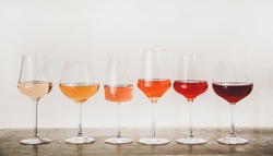 Various shades of Rose wine in stemmed glasses placed in line from light to dark colour on concrete table, white wall background behind. Wine bar, wine shop, wine tasting concept