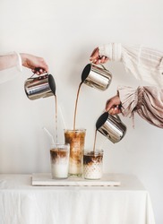Female hands in elegant dress sleeves pouring fresh coffee to glasses with milk and making latte cappuccino drinks for breakfast on board over light wall background. Coffee beverage drinks