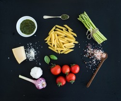 Ingredients for cooking pasta. Penne, green asparagus, basil, pesto sauce, garlic, spices, parmesan cheese and  cherry-tomatoes on dark grunge backdrop, top view