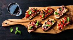 Brushetta set for wine. Variety of small sandwiches with prosciutto, tomatoes, parmesan cheese, fresh basil and balsamic creme served with glass of red wine on rustic wooden board over dark background
