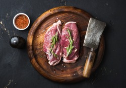 Raw uncooked poultry meat cut. Duck breast with rosemary, spices and butcher cleaver on dark wooden tray over black wooden background, top view