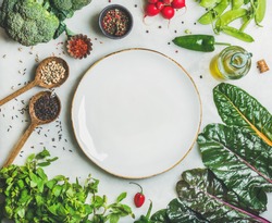 Fresh raw greens, vegetables and grains over light grey marble kitchen countertop, wtite ceramic plate in center, top view, copy space. Healthy, clean eating, vegan, detox, dieting food concept