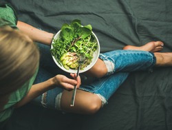 Green vegan breakfast meal in bowl with spinach, arugula, avocado, seeds and sprouts. Girl in jeans holding fork with knees and hands visible, top view. Clean eating, detox, vegetarian food concept