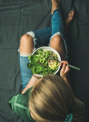 Green vegan breakfast meal in bowl with spinach, arugula, avocado, seeds and sprouts. Girl in jeans holding fork with knees and hands visible, top view. Clean eating, dieting, vegetarian food concept