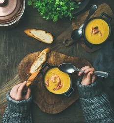 Woman in sweater eating corn creamy soup with shrimps in individual pot, top view. Woman' s hand keeping spoon and bread slice. Flat-lay of rustic dinner table. Slow food, winter warming food concept