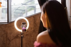 Young woman making photo or video content for social media with smartphone and light of ring lamp. Beauty blogger smiles to mobile phone screen. Influencer Makes broadcast communicates with followers