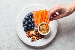 Woman hand eating healthy snacks plate - carrots, nuts, berries and peanut butter, top view.