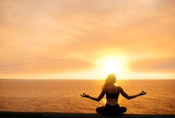 A girl practices yoga by the sea during a beautiful sunset. She is  sitting in lotus position and meditating