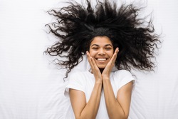 Top view portrait of joyful african american woman with curly hair, is lying on the bed, looks directly at the camera and smiling. Beautiful female enjoy weekends at home