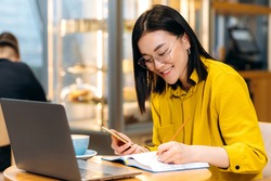 Modern Asian female student in stylish casual clothes, studying remotely using a laptop, listening to an online lesson, taking notes in a notebook, while sitting in a coffee shop. Online education