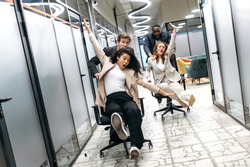 Funny young adult business people making a race on chairs in modern office. Multiracial friendly colleagues spending together their work break, resting from brainstorm