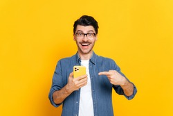 Happy joyful caucasian stylish guy with glasses, holding smartphone in hand and pointing at it finger, looking happily at camera with smiling, standing on isolated orange background