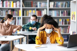 A tired African-American female student in a protective medical mask sits at a table in a library with a laptop, eyes closed, thinks. In the background,two students greet each other with an elbow bump