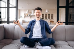 Relaxed attractive young caucasian man, calm pacified guy in casual clothes sits on the sofa in the living room with closed eyes, meditates in the lotus position, relieves stress, resting