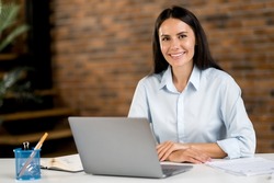 Portrait of a confident successful young adult caucasian brunette business woman, ceo, broker, manager sitting in office at table, using laptop, in formal stylish clothes, looking at camera, smiling