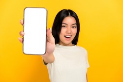 Lovely excited chinese positive brunette young woman in casual t-shirt showing modern smart phone with blank white screen, standing on isolated yellow background, smiling. Mockup, copy space