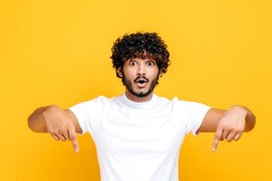 Positive indian or arabian happy guy in basic white t-shirt, amazed looks at camera and points fingers down, surprised face expression, stands on isolated orange color background