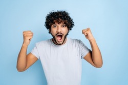 Enthusiastic amazed indian or arabic guy, in abasic t-shirt, rejoices in success, victory, win, gesturing with fists, looking at the camera, smiling, standing on an isolated blue background