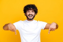 Amazed indian or arabian cheerful young man in white basic t-shirt looks at the camera and points fingers down at space for your presentation, stands on isolated orange color background, smiling