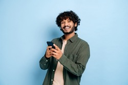 Happy handsome amazed stylish indian or arabian guy, using smartphone, chatting online, texting message, browsing internet, social media, looks at camera, stands on isolated blue background, smiling