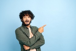 Surprised shocked indian or arabian guy in casual clothes, amazed looks at camera and points finger to the side at space for presentation, stands on isolated blue background, smile. Copy space concept