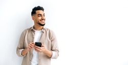 Panoramic photo of a joyful handsome indian or arabian guy, holding smartphone in hand, chatting online, browsing internet, looking happily to the side, standing on isolated white background, smiling