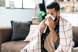 Sneezing, runny nose, cough. Ill arabian or indian young man, sits on the couch at home under a plaid, sneezing in a napkin, feels weak, unwell, needs treatment and medicines