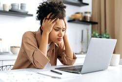 Desperate worried sad african american young woman, using laptop for work or study from home, sitting in kitchen, frustrated looking at screen, failed project, got financial loss, job downgrade