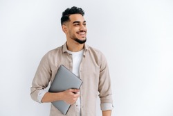 Cheerful arabian or indian young man with beard, wearing casual shirt, standing over isolated white background, holding laptop, looking to the side, smiling joyfully, dreaming, thinking