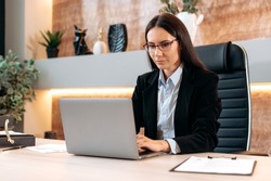 Intelligent caucasian beautiful brunette woman with glasses, in a formal suit, top manager, realtor, broker, sits at a desk in modern office, works in a laptop, chatting online with clients