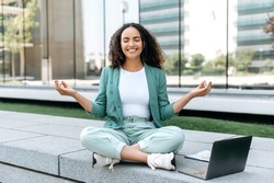 Calm relaxed curly haired latino or brazilian woman, company worker, sits in lotus position outdoors near the business center, takes break during work time, meditates, relaxes, closed her eyes, smiles
