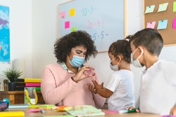 Teacher cleaning hands to student children with sanitizer gel while wearing face mask in preschool classroom during corona virus pandemic - Healthcare and education concept