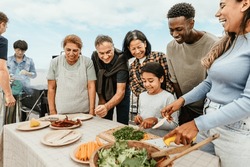 Multi generational people having fun doing barbecue at house rooftop - Happy multiracial friends cooking together - Summer gatherings and food concept
