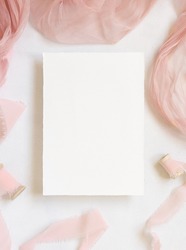 Paper card on white marble table with dried pink flowers and silk ribbons top view. Flat lay with vertical blank card. Romantic invitation or greeting card mockup, copy space