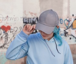 Blue haired Teenage girl in blue hoodie and baseball cap touching her cap. Blue haired teen girl stays outdoors against graffiti wall. Clothing and cap mockup 