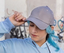 Blue haired Teenage girl in blue hoodie and baseball cap with closed eyes touching her cap. Blue haired teen girl stays outdoors against graffiti wall. Cap mockup 