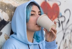 Teenage girl in light blue oversize hoodie drinking coffee to go. Blue haired teen girl outdoors near graffiti wall. Coffee cup mockup. Hipster and adolescence concept. Head shot