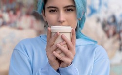 Teenage girl in light blue oversize hoodie with coffee to go and looking stright.Blue haired teen girl staying outdoors near graffiti wall, head shot.  Hipster and adolescence concept