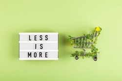 Less is more - written on lightbox next to Shopping cart entwined with plants on green background. Conscious consumption slow fashion Zero waste concept. Top view flat lay