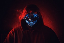 Man in angry and scary lighting neon glow mask in hood on dark red background. Halloween and horror concept.