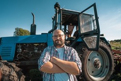 Portrait of satisfied tractor driver after work on agricultural field stands next to tractor. Farming and harvesting