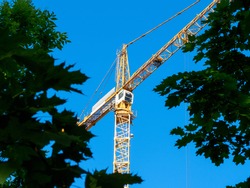 low angle view of a construction tower crane through green foliage. Funds for building construction
