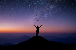 Silhouette of young woman standing alone on top of mountain and raise both arms praying and free bird enjoying nature on beautiful night sky, star, milky way background. Demonstrates hope and freedom.