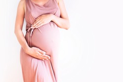 Pregnant woman in dress holds hands on belly on a white background. Pregnancy, maternity, preparation and expectation concept. Close-up, copy space. Beautiful tender mood photo of pregnancy.